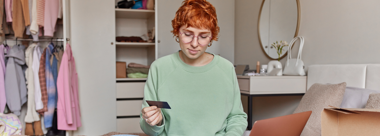 Student zit op haar kamer met allerlei pakketjes om haar heen en heeft een bankpas in haar hand en checkt haar saldo op haar latop
