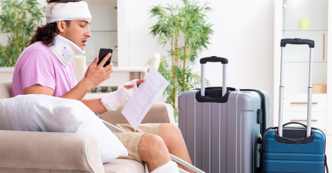 Jonge man met nekkraag, krukken, verband om zijn hoofd en been zit op de bank met zijn telefoon en een papier in zijn hand naast zijn koffers